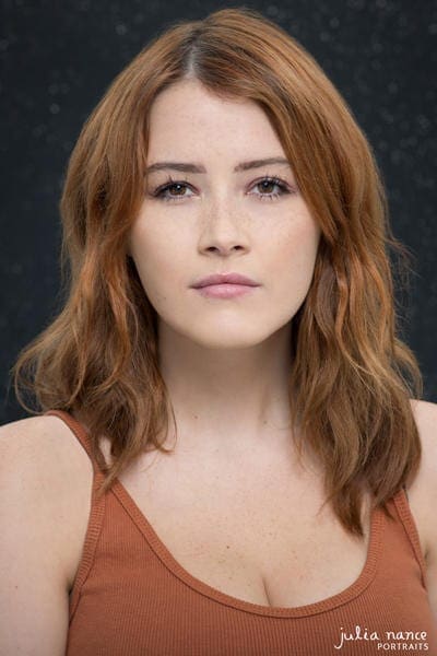 Melbourne Actor Headshot of redhead woman with a burn orange top on. She has a moody expression and the photograph has a dark background.