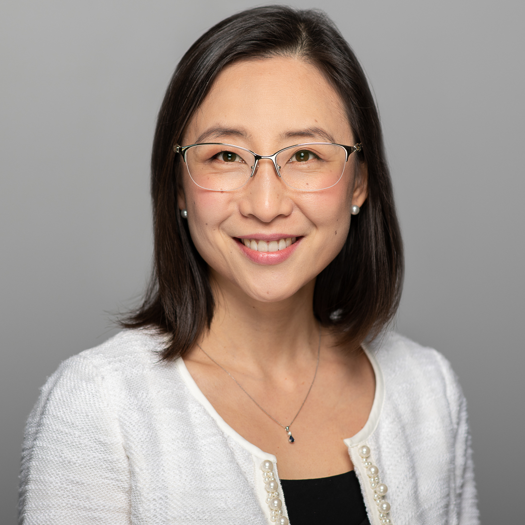 Studio corporate headshot of woman wearing white cardigan and glasses, smiling at the camera.