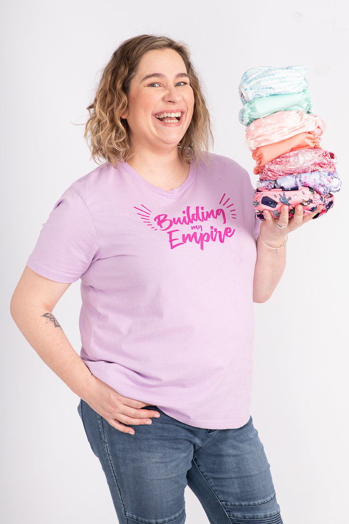 Woman holds a collection of cloth nappys while her tshirt reads "building my empire". She is smiling at the camera.