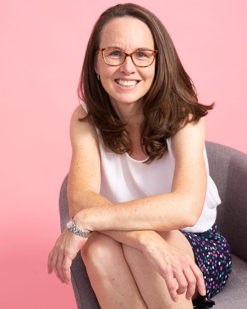 Woman smiles to camera seated in an armchair on a pink background