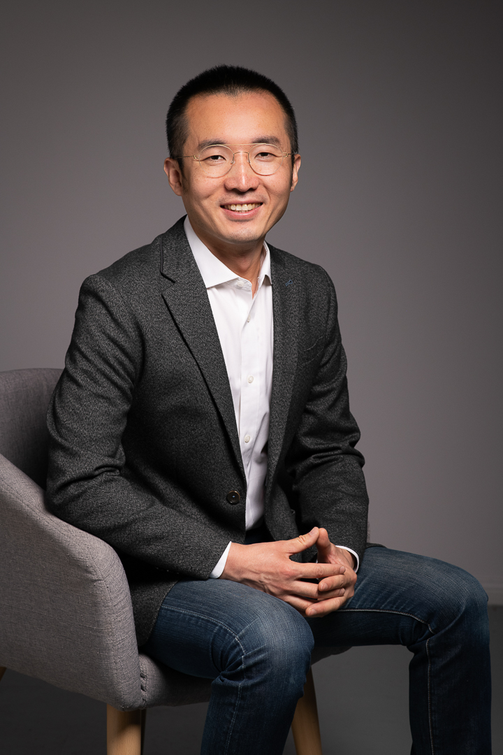 Corporate headshot of man seated in an armchar. He has his hands together and wears a professional blazer, top and jeans.