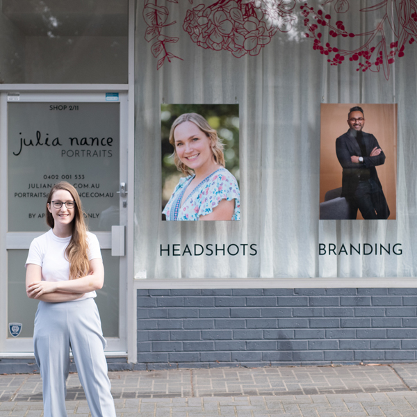 Julia Nance stands out the front of her professional portrait studio. She has blonde hair, glasses and has her arms crossed in a relaxed manner.