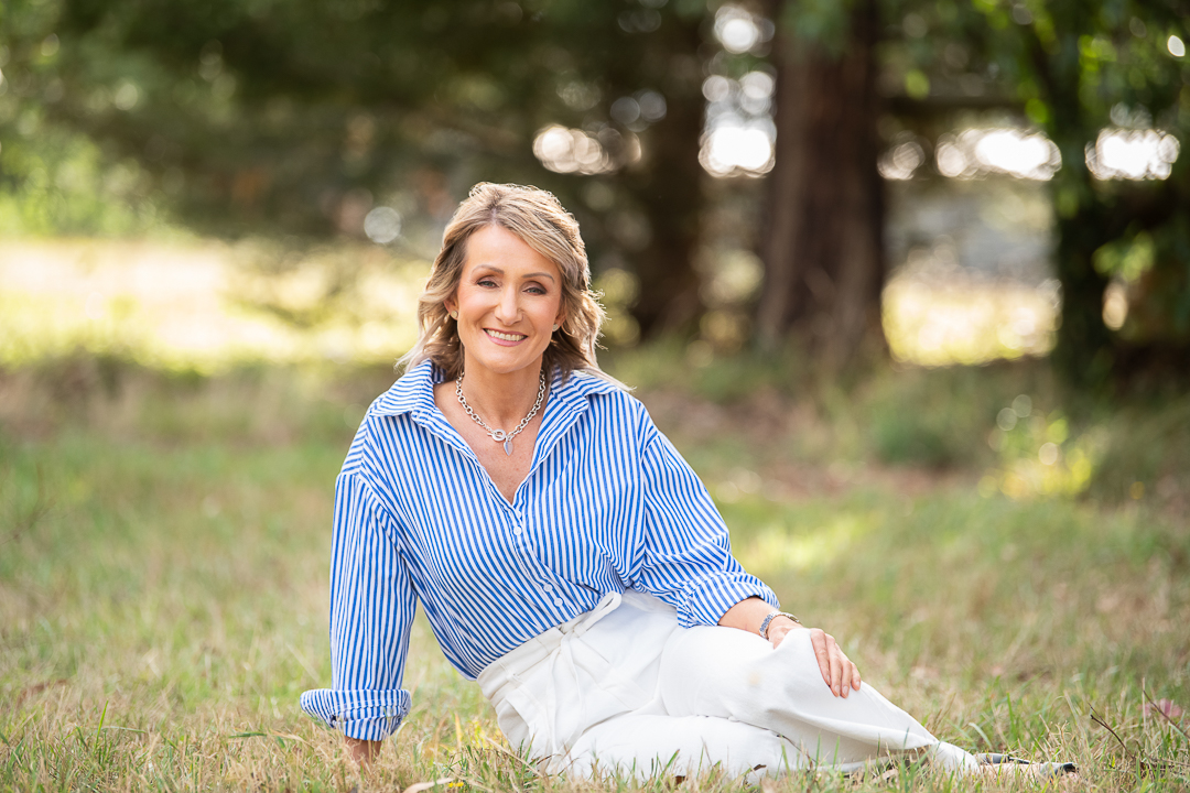 Relaxed personal branding portrait of woman sitting outside on grass, smiling to camera.