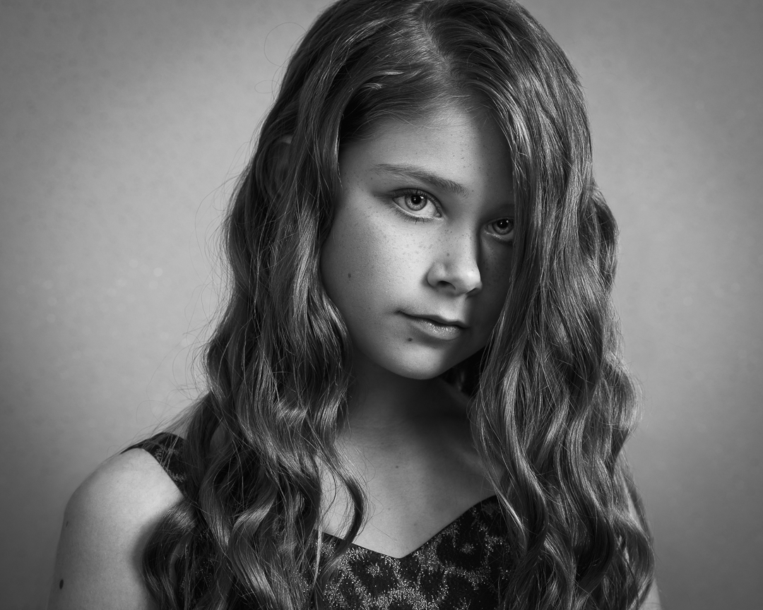 Fine art model headshot of teen model in black and white. Girl looks directly to camera with curled hair falling over one half of her face.