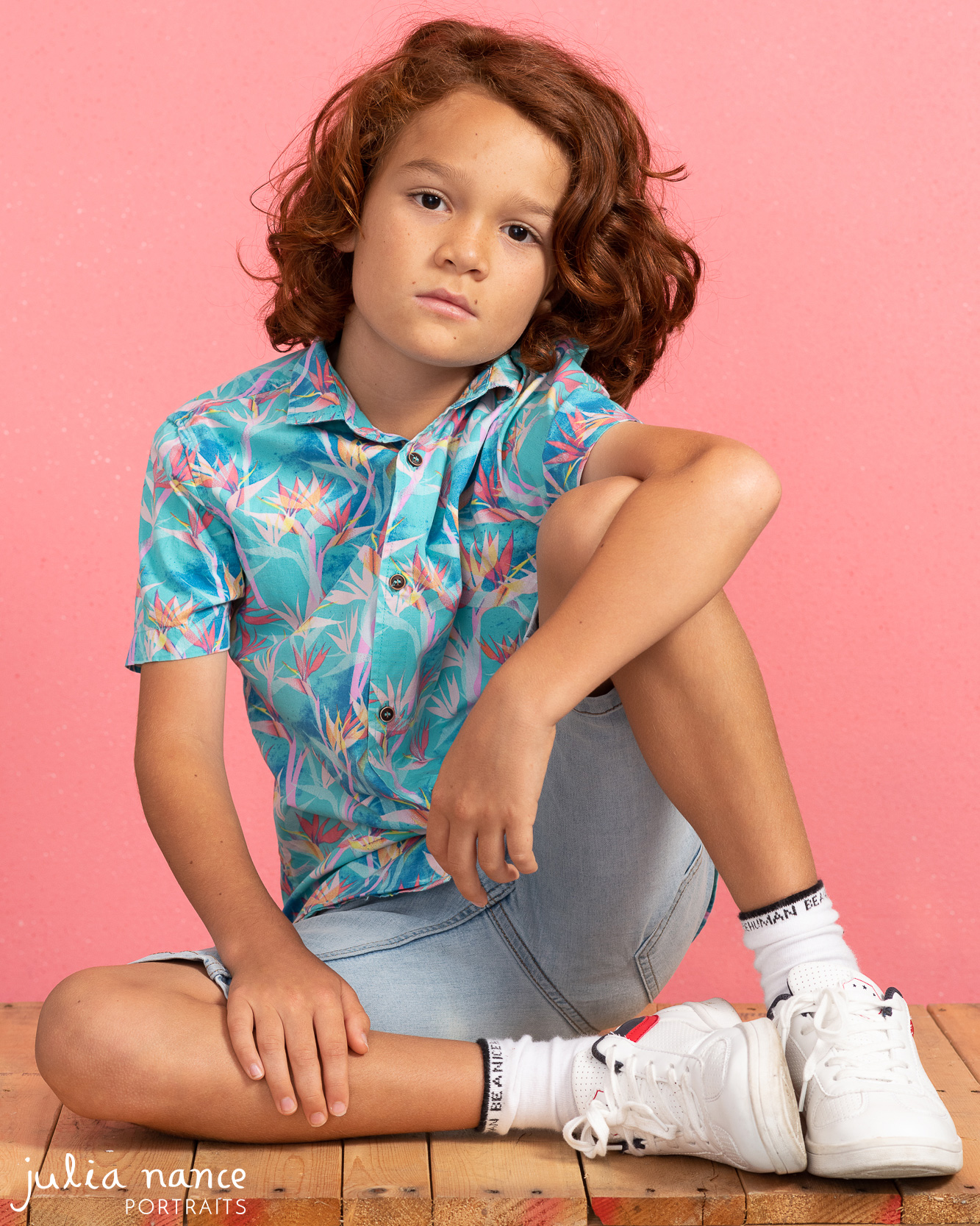 Pre-teen boy sits in front of a pink background wearing a blue floral shirt with a serious expression for his model portfolio photoshoot in Melbourne.