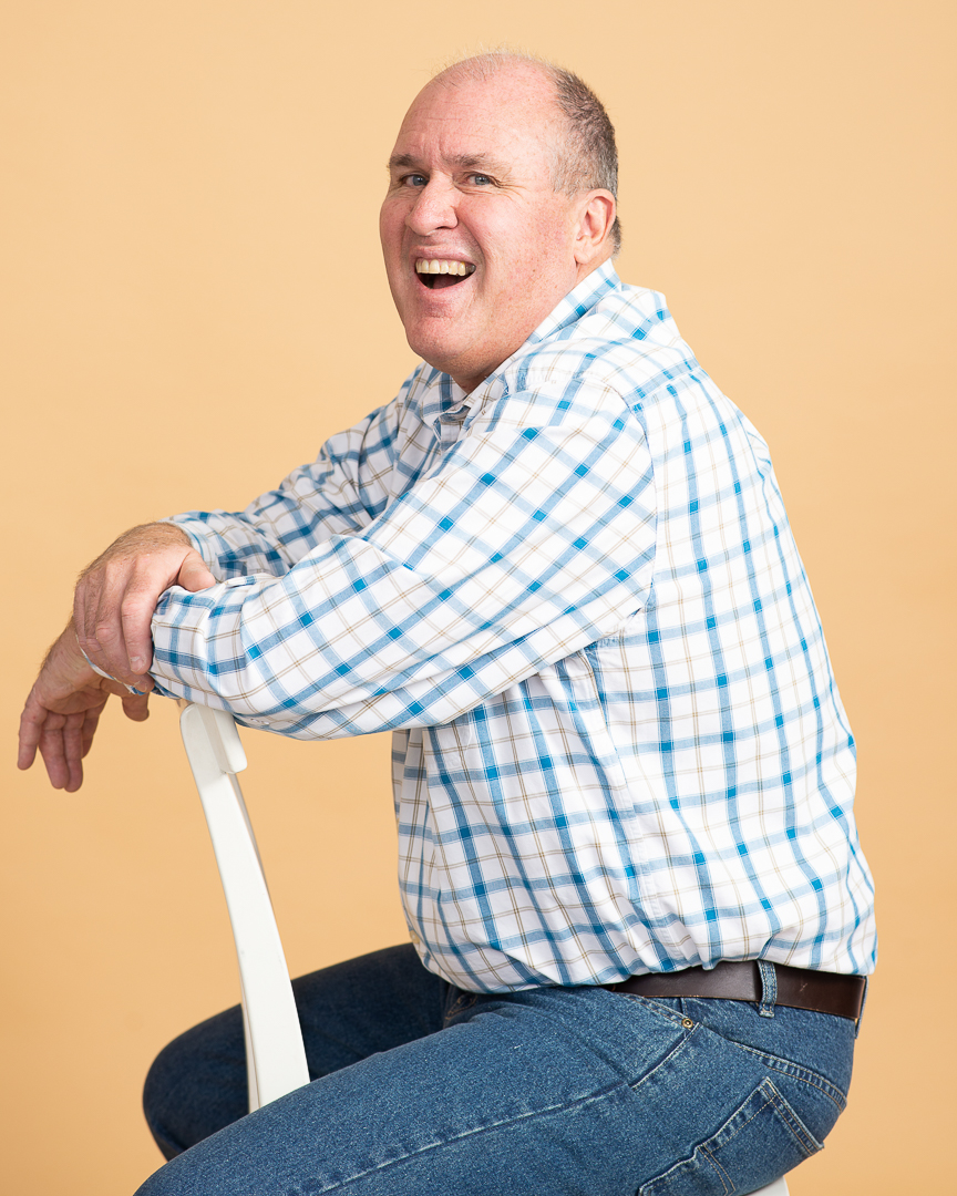 Melbourne man sits on a white chair leaning on the back. He laughs to camera with a joyful smile, while wearing a checkered shirt and jeans.