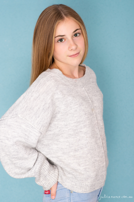 Teen girl poses for a relaxed model portfolio headshot in front of a blue background.