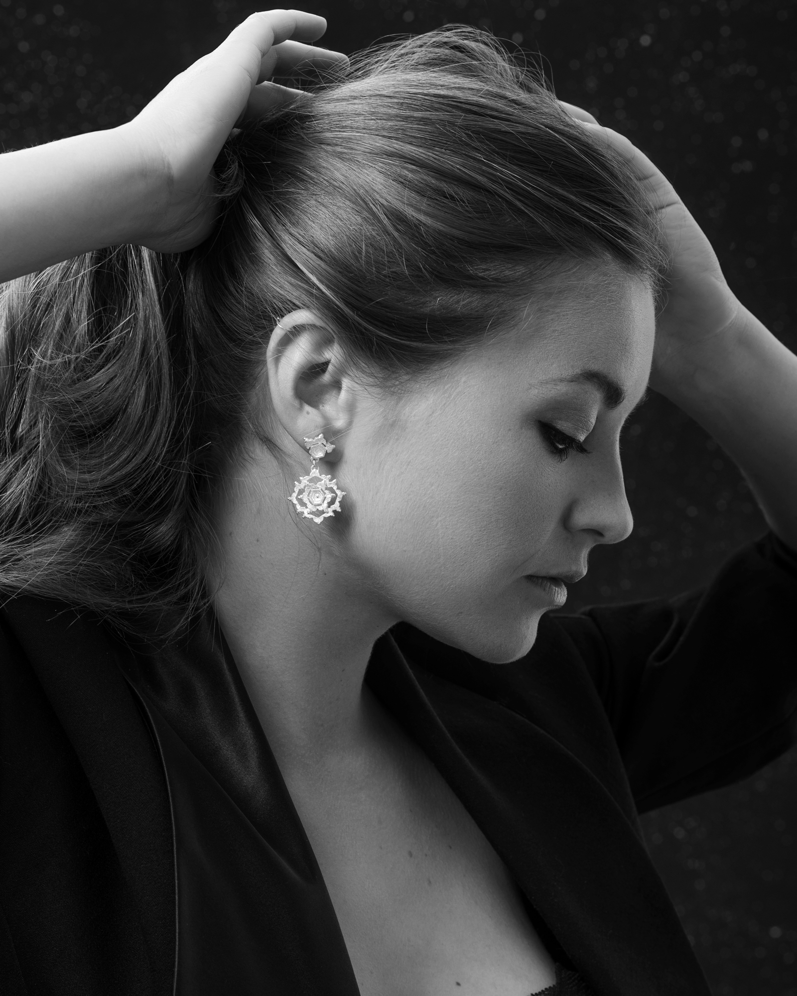A beauty headshot of model. Image is close up showing model's side profile. she has her hands up and pulls her hair back while looking down. The image is in black and white.