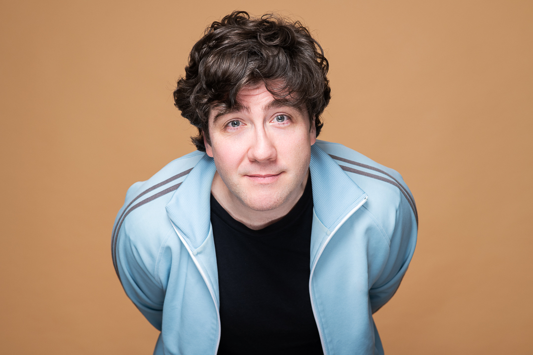 Melbourne actor and comedian pete in a headshot update. He leans right in to camera with a quirky smile, wearing a blue jacket on an orange background.