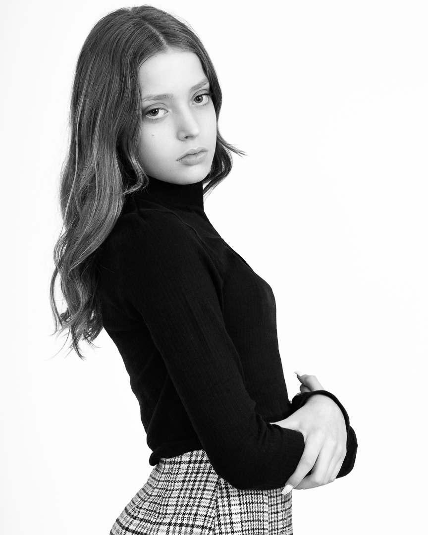 A model portfolio portrait of girl on white background wearing a black turtleneck top and skirt. She looks over her shoulder with a serious expression.