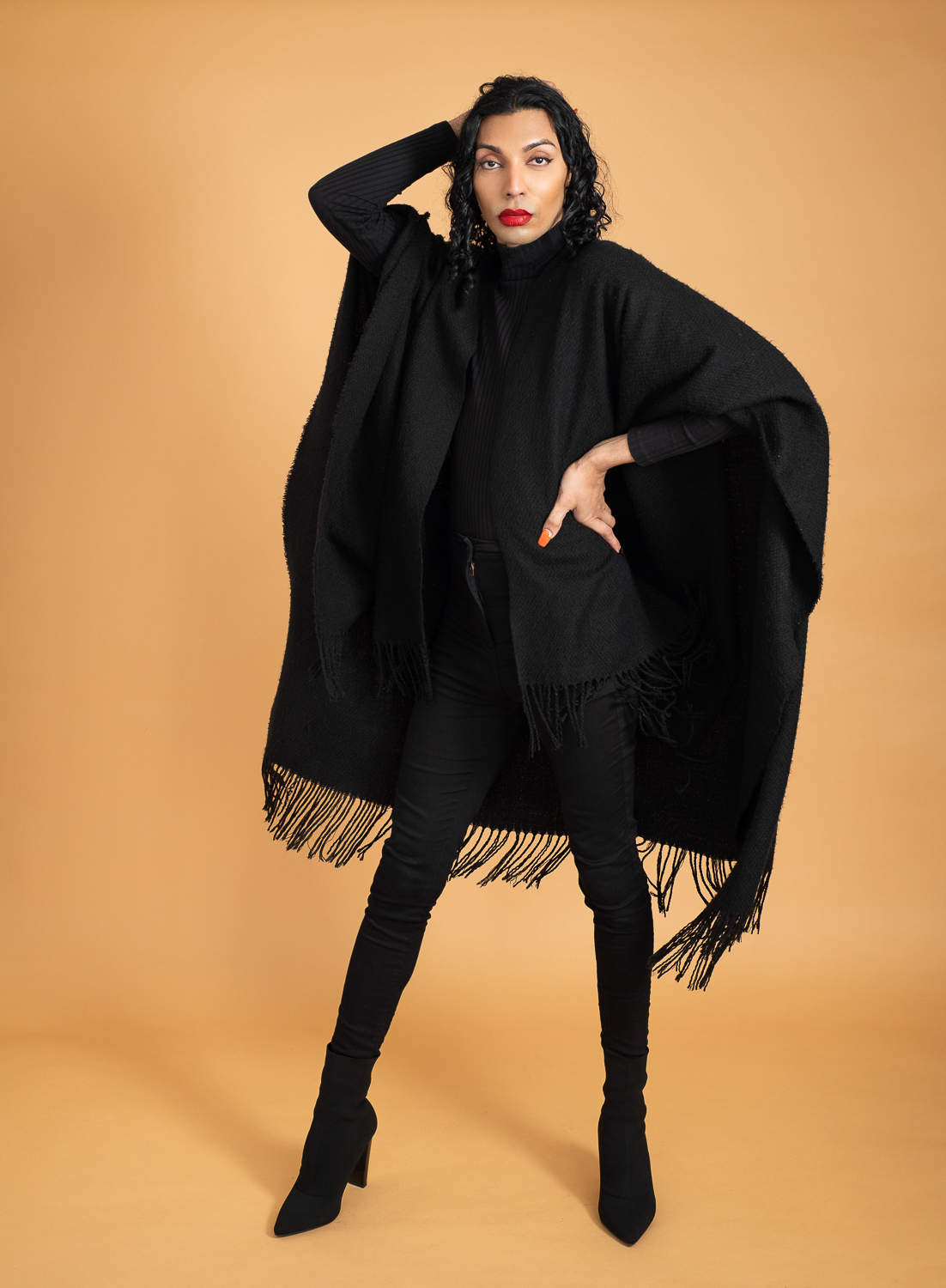 Melbourne trans model poses in an all black outfit on an orange seamless background. They have a confident pose and expression and wear red lipstick.