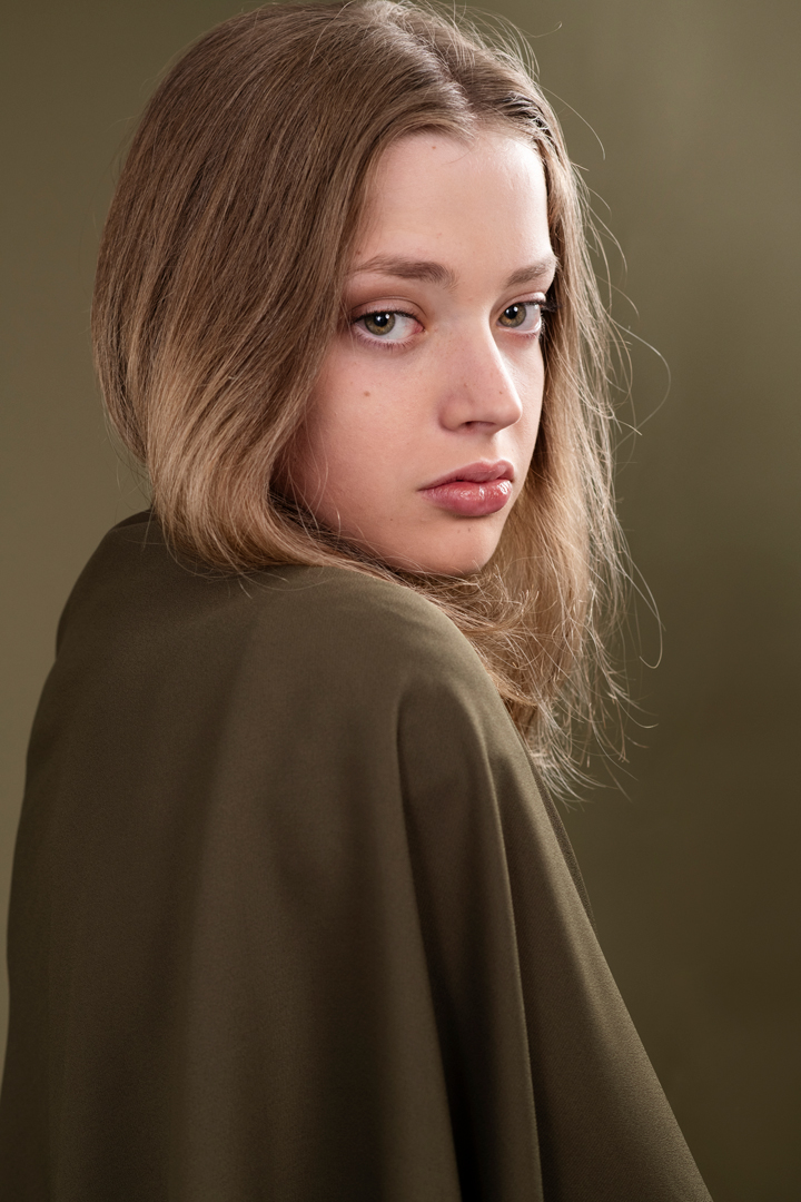 Teen model looks over her shoulder in a monochromatic olive green fashion portrait. She has green eyes and blond hair, for a striking model headshot.
