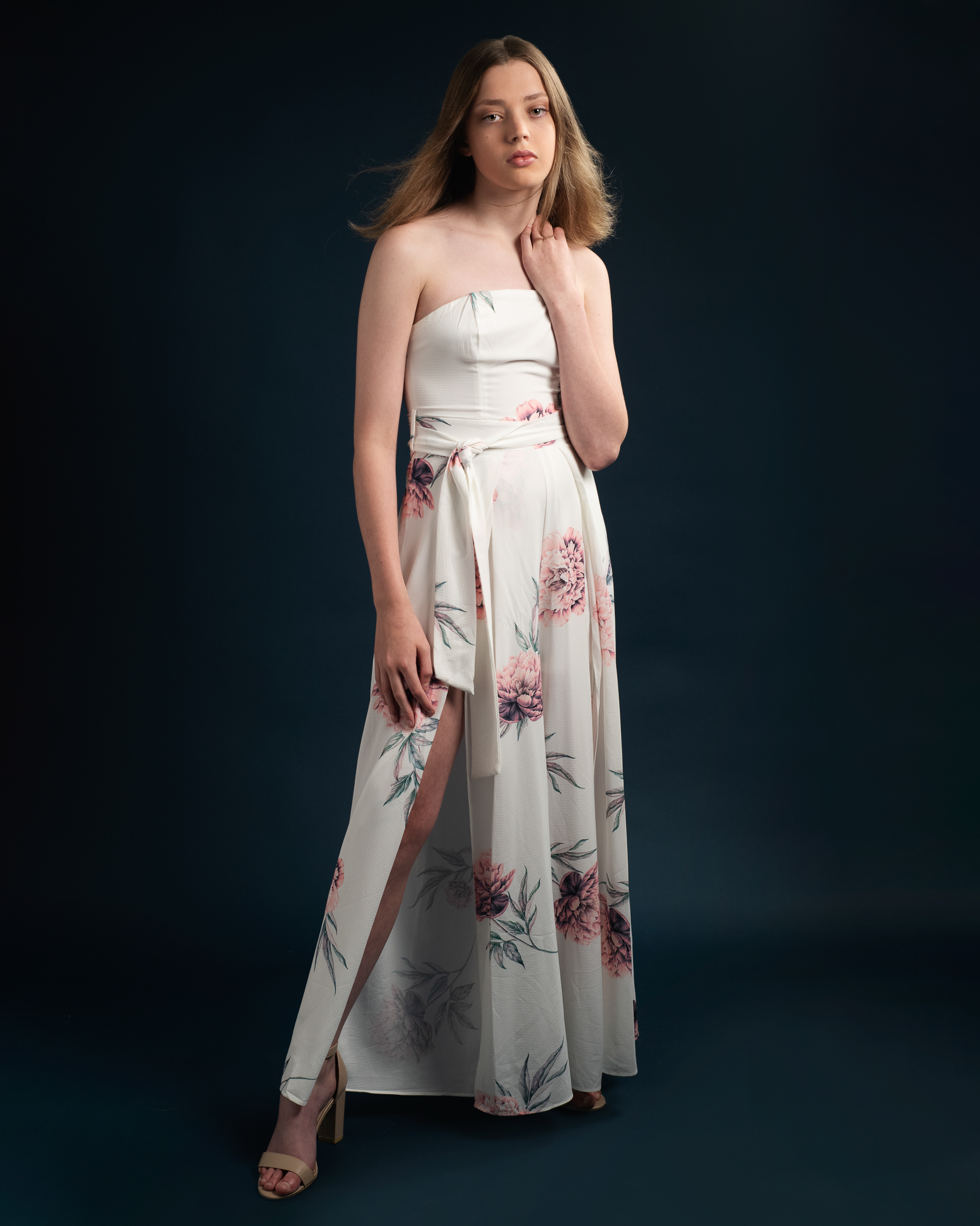 Teen model stands on navy background in Melbourne studio posing to the camera in a full length image. She wears a white floral dress and looks at the camera with a serious expression while wind blows her hair.