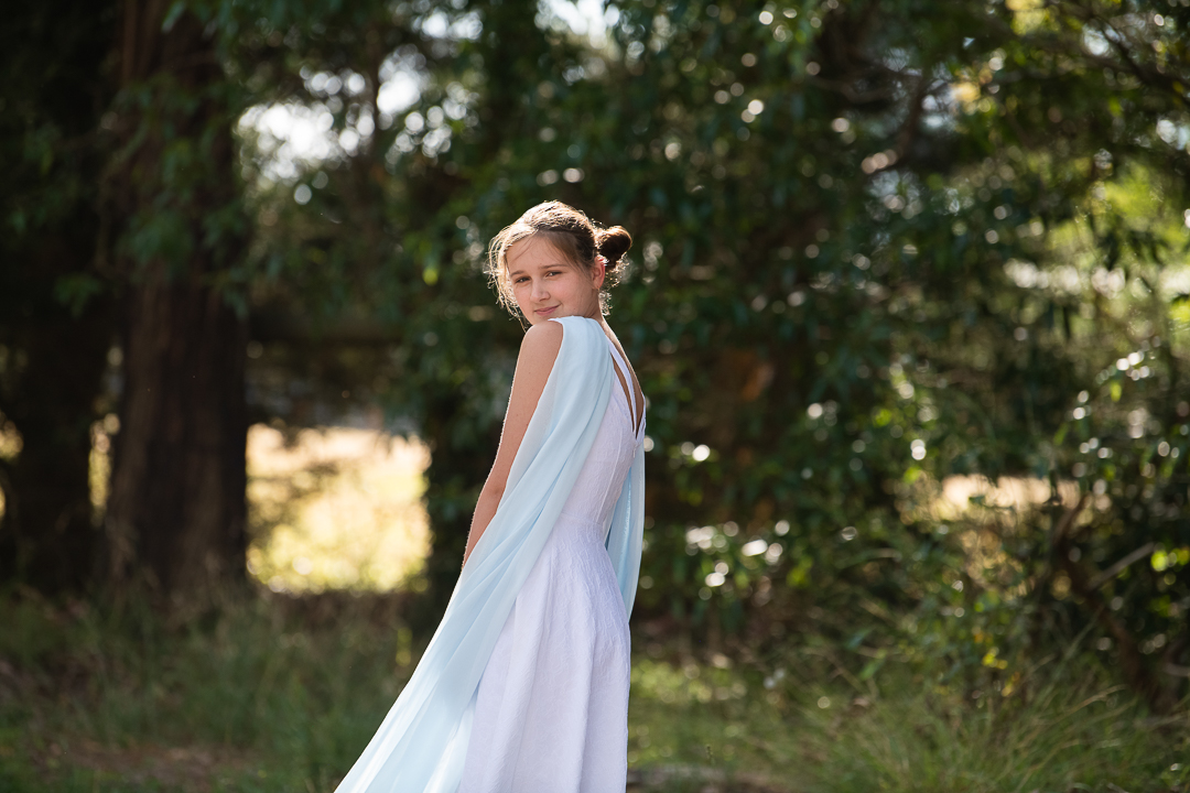 Young teen model and actor poses outdoors for a model portfolio shoot in the Melbourne sunshine. She wears a white dress with a pale blue scarf, blowing in the wind.