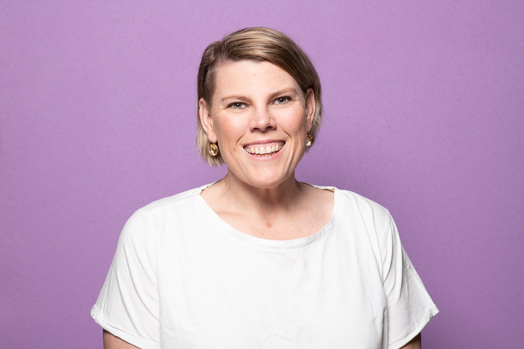 A bright and happy headshot of melbourne woman who smiles to camera on a bright purple background.