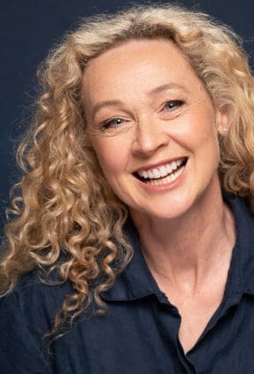 Melbourne actress angela laughs to camera in a studio actor headshot. She wears a navy collared shirt and is photographed on a navy background.