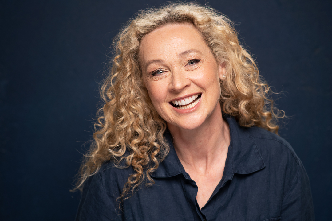 Melbourne actress angela laughs to camera in a studio actor headshot. She wears a navy collared shirt and is photographed on a navy background.