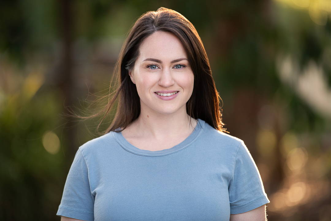 Natural outdoor actor headshot taken at golden hour in melbourne's eastern suburbs. Woman wears a pale blue tshirt and smiles to camera with the wind blowing through her hair.