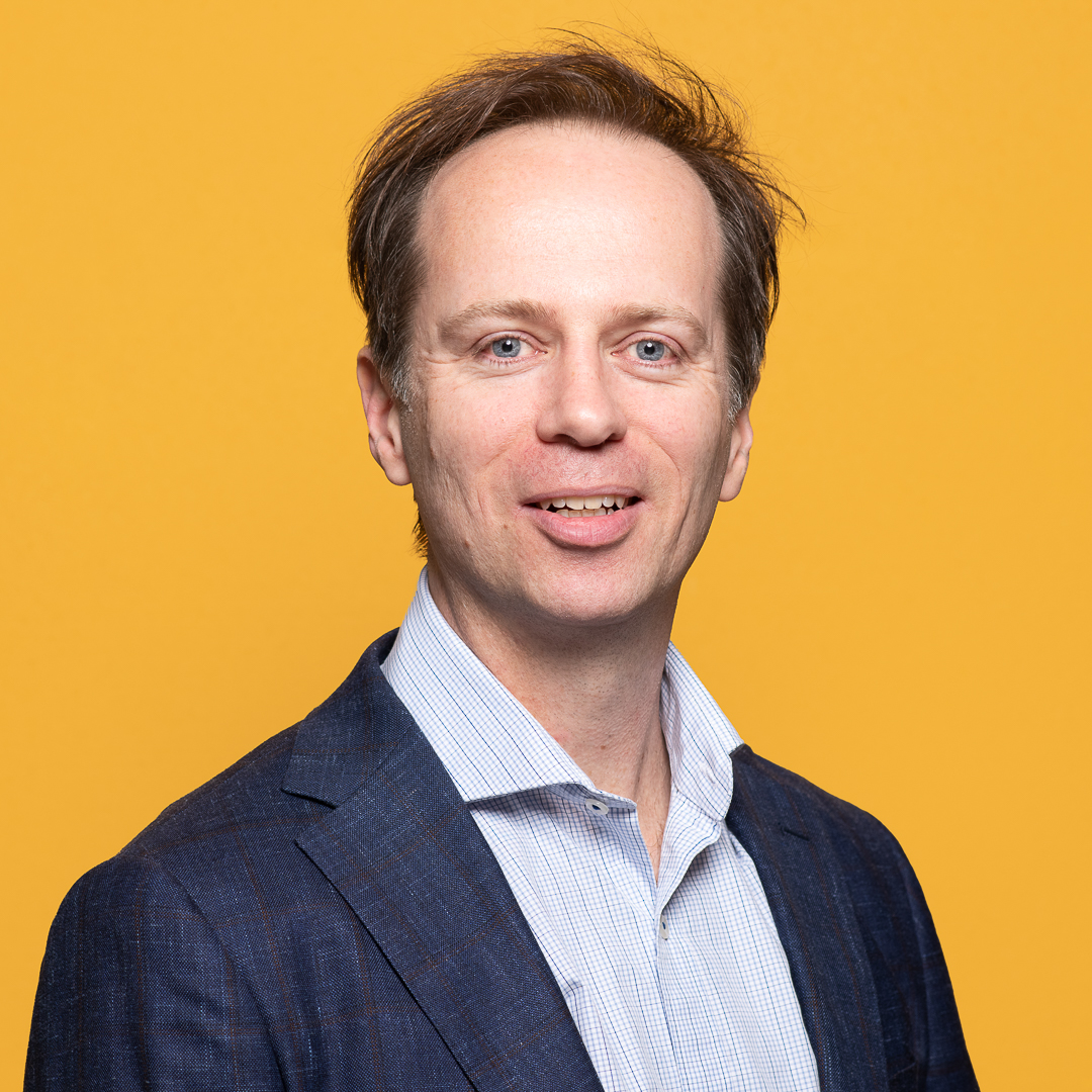 A studio portrait of melbourne professional for a new linkedin headshot. There is a bright yellow background behind him and he smiles to camera.