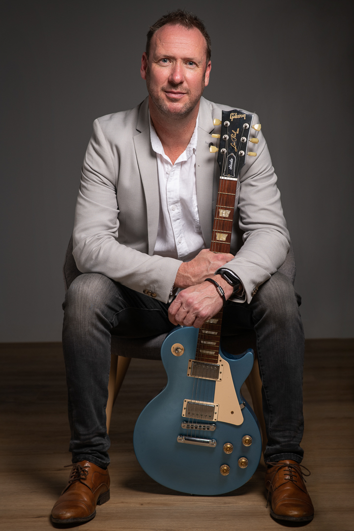 Melbourne musician glen holds a blue electric guitar while seated for a professional branding portrait in studio.