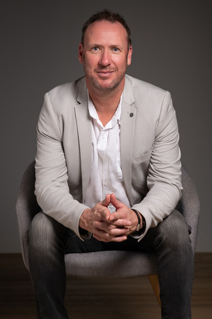Melbourne musician Glenn is seated in a grey armchair in a portrait studio, wearing a professional suit. He leans on his knees in a professional poses and smiles to camera.