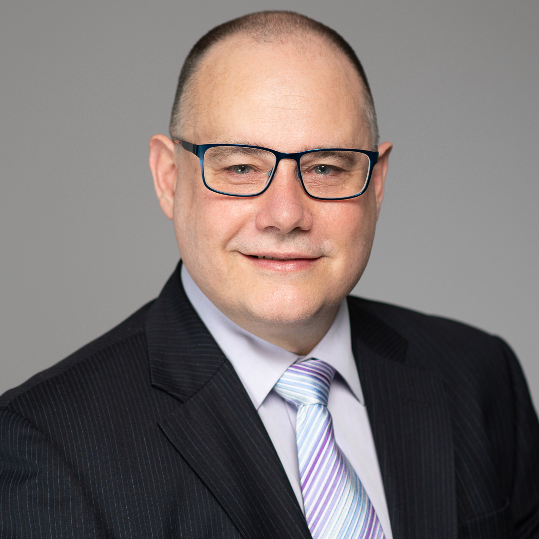 Professional corporate headshot of melbourne accountant wearing glasses and a formal suit. He smiles to camera on a light grey studio background.