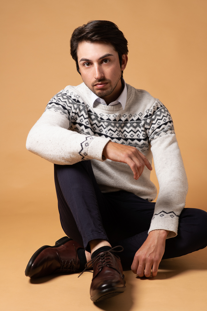 Relaxed model portrait of male model on earthy orange background wearing a casual jumper and dark pants, while seated on a seamless studio background.