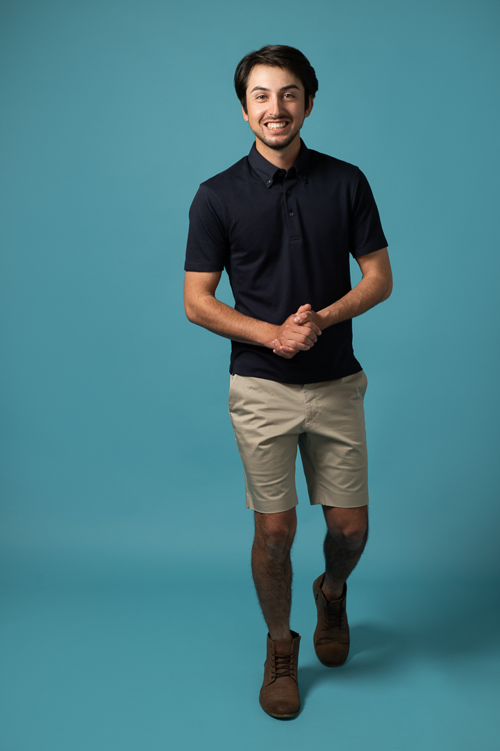 Melbourne male model smiles to camera while walking on a blue background with his hands together in a relaxed portrait pose.