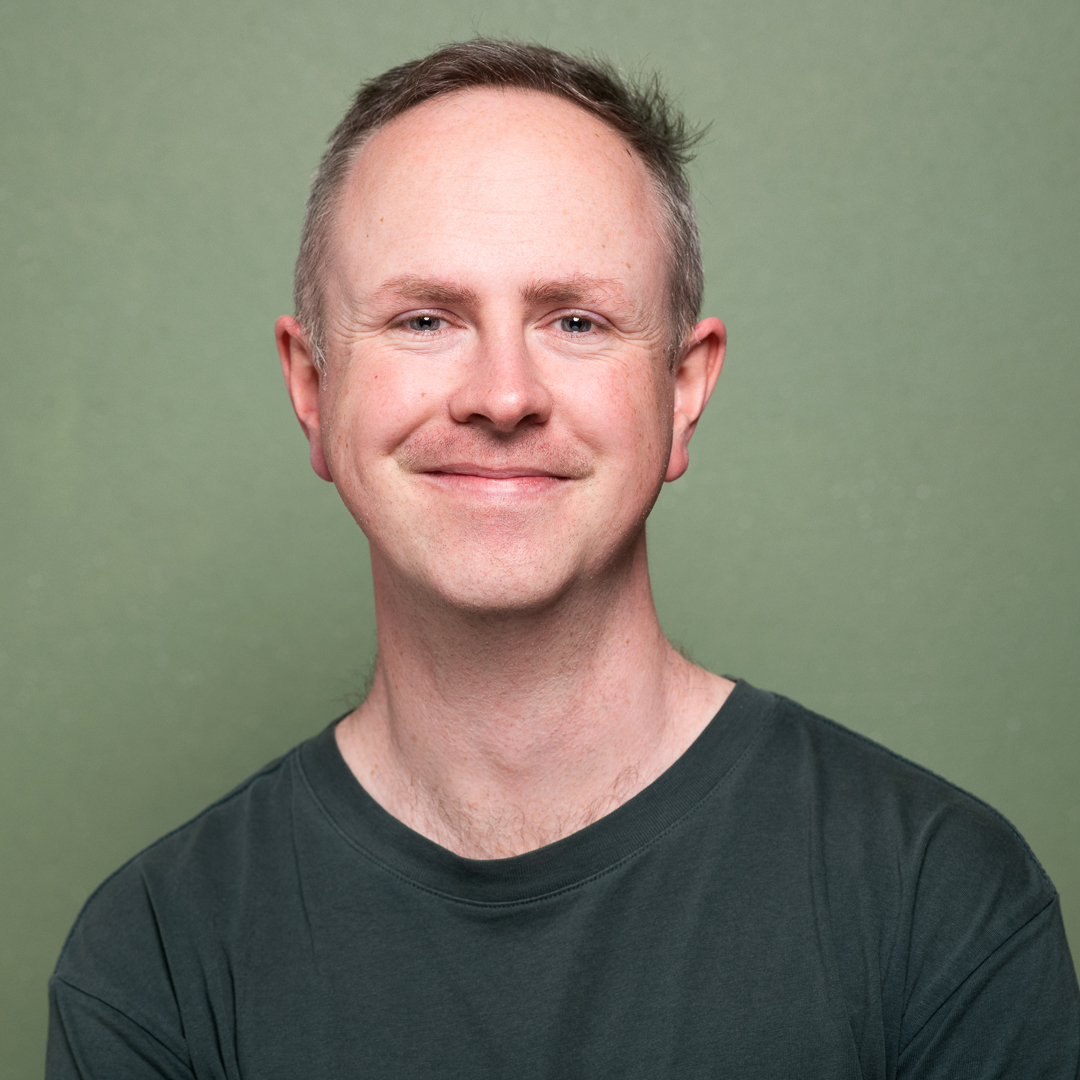 A melbourne professional smiles to camera wearing a green tshirt for a linkedin headshot. It was taken on a green background in a melbourne portrait studio.