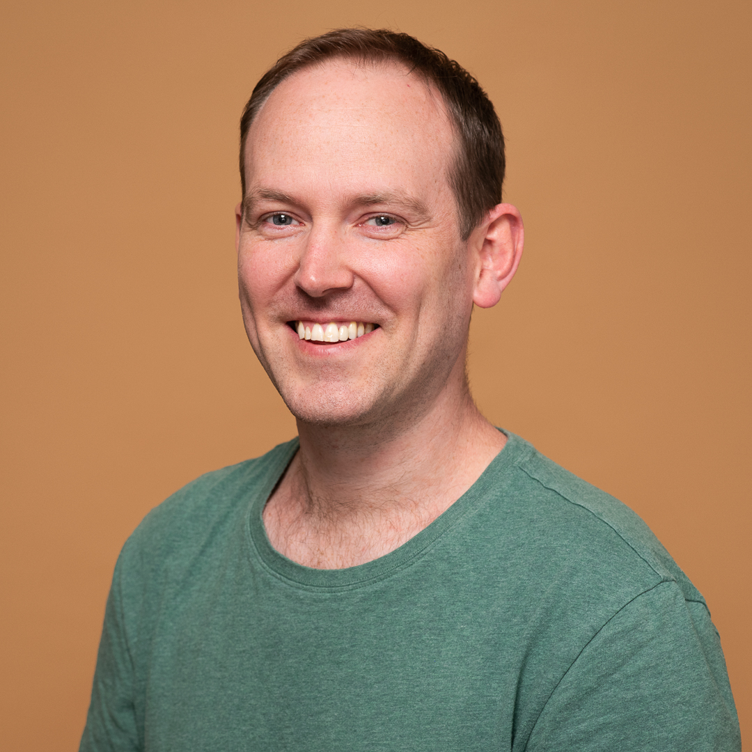 A relaxed linkedin headshot of melbourne man wearing a green tshirt with an orange background behind him. He has a natural smile and looks at the camera.