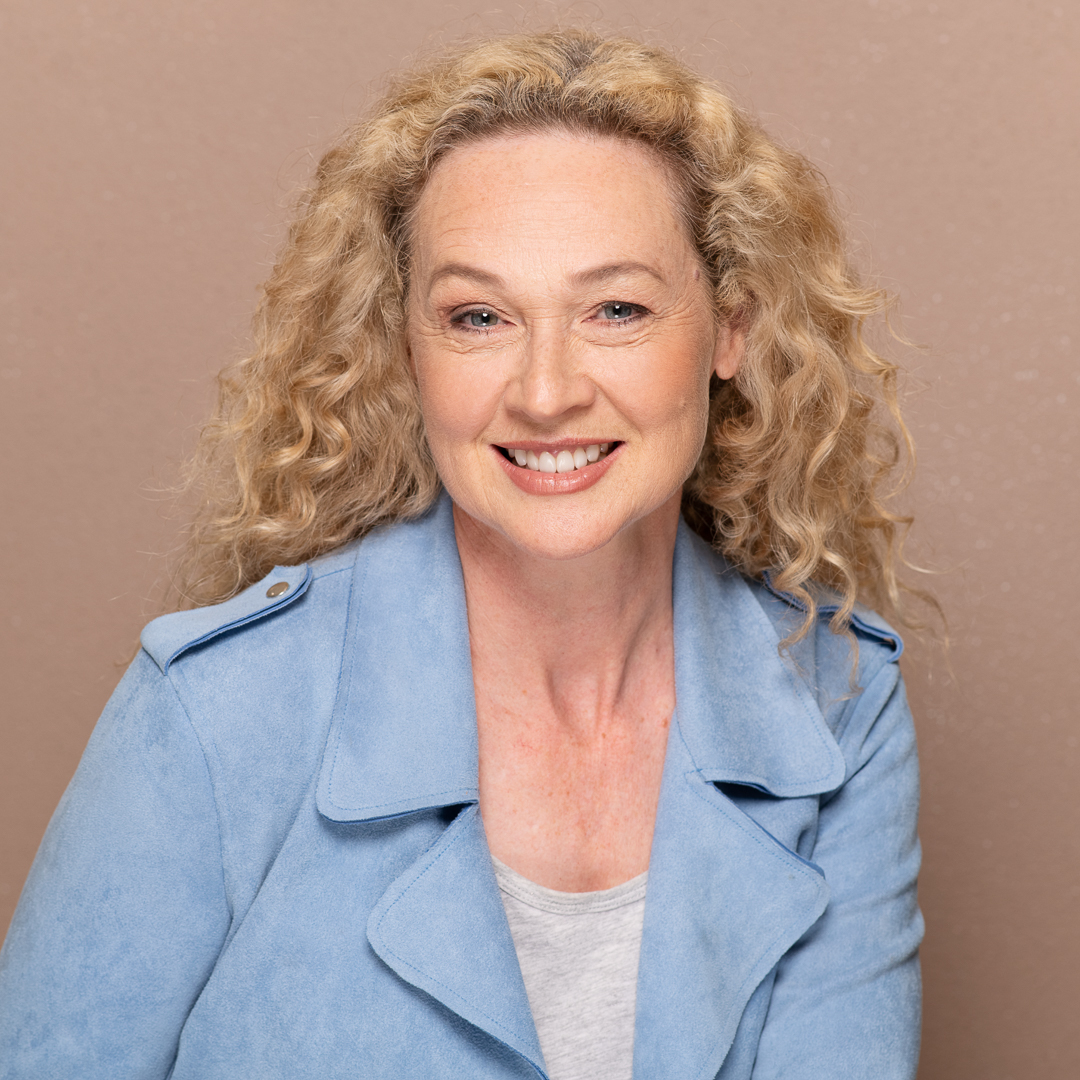 Melbourne actress smiling for a relaxed actor headshot. She wears a blue blazer and is in front of an earthy brown background in a studio.