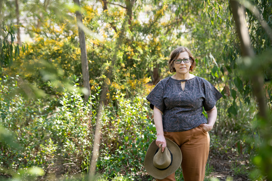 Model portrait of mature age model in an outdoor melbourne landscape filled with trees and wattle. She has one hand in her pocket and is holding her hat in the other, smiling to camera.