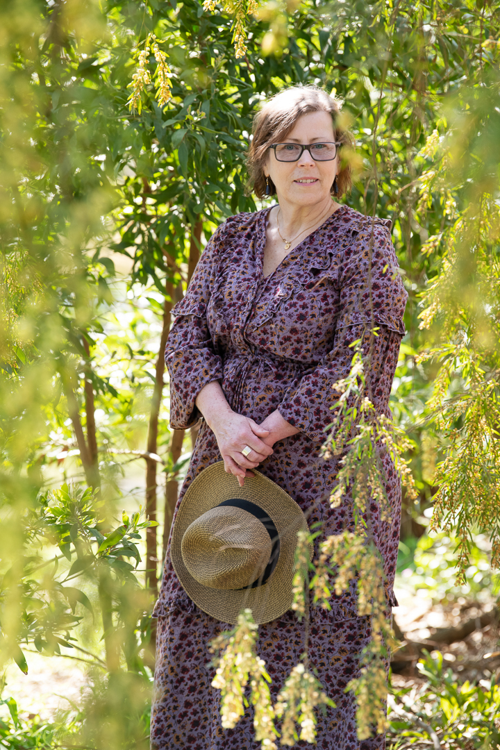 Model portrait of woman in an outdoor landscape, surrounded by yellow wattle, taken in Melbourne's eastern suburbs. She wears a purple dress and is holding a hat.