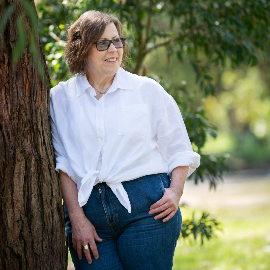Mature model portrait of woman outdoors leaning against a tree. She looks away from the camera wearing glasses, jeans and a white linen shirt.