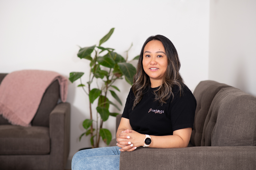 A personal branding portrait of melbourne mortgage broker seated on a couch with a green plant behind her. She wears a branded tshirt, jeans and heels.