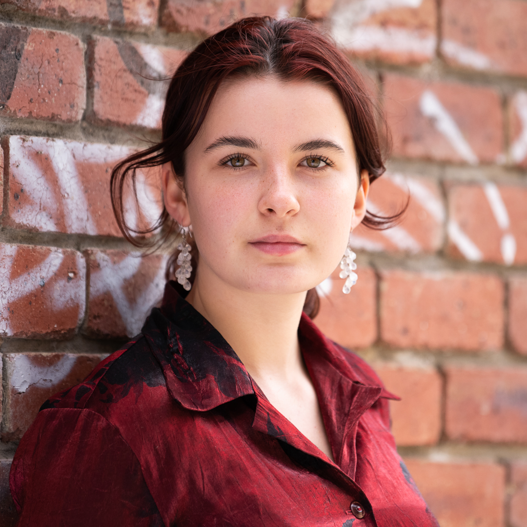 Actor headshot of young melbourne actress looking directly to camera. She has red hair and a deep red blouse, standing next to a brick wall.