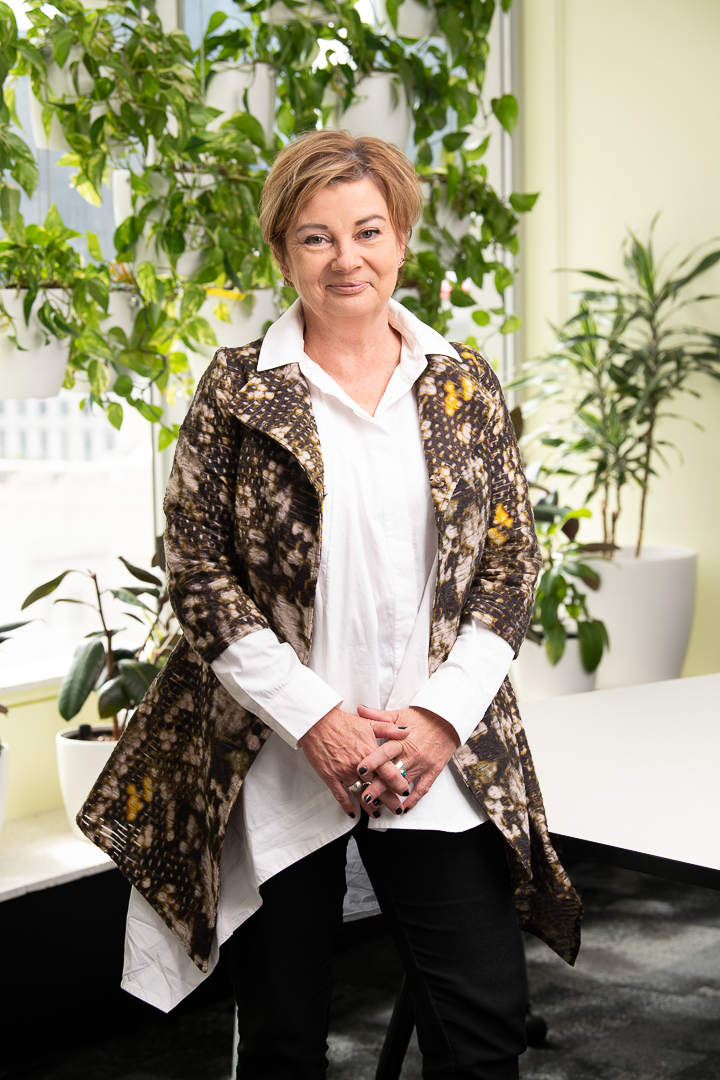 Staff headshot of woman smiling and standing in office with melbourne city out the window and indoor plants filling the space.