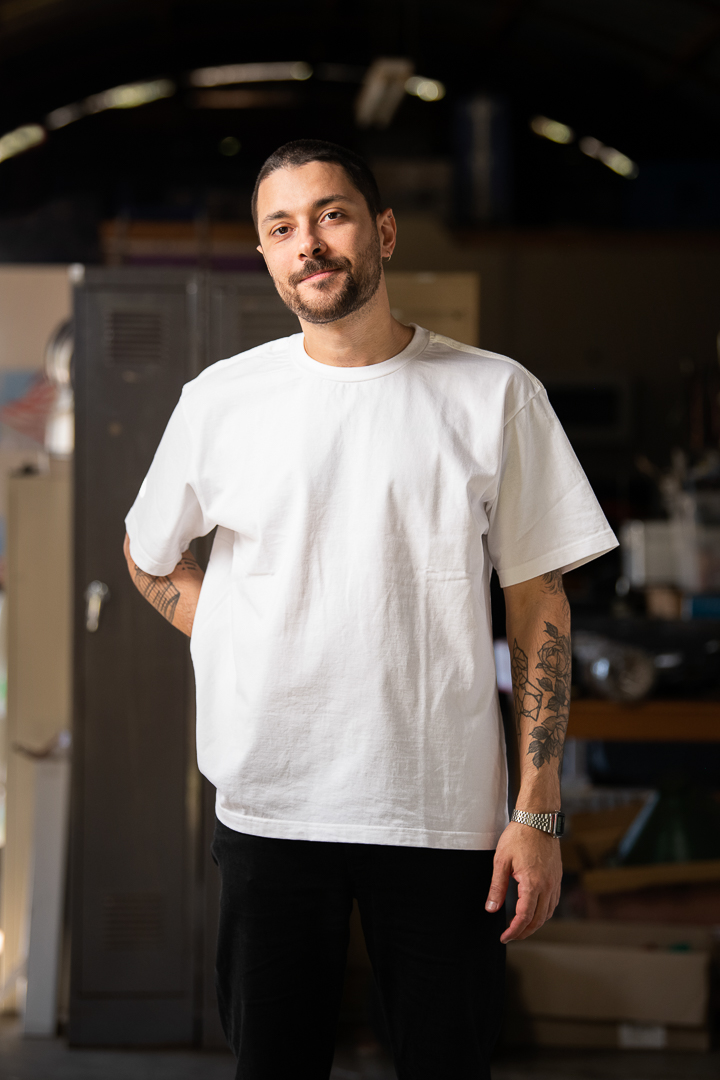 A personal branding portrait of melbourne man Jean, who wears a white tshirt in a warehouse setting. He smiles to camera and stands confidently.