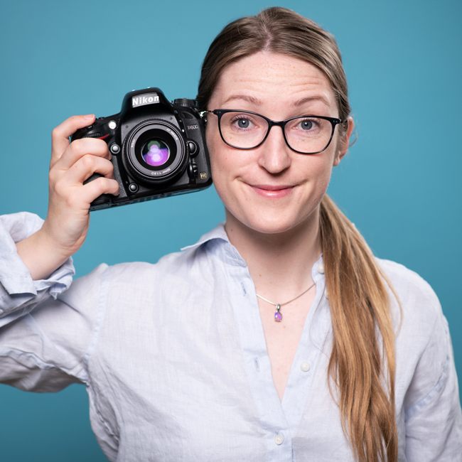 Professional headshot of Julia nance holding a camera next to her cheek, smiling to camera on a blue background.