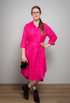 Portrait of Julia Nance wearing a bright pink dress with one hand on her hip and the other holding her camera. She smiles to camera and stands confidently.