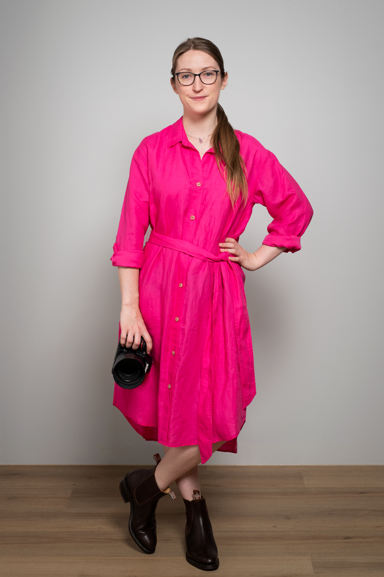 Portrait of Julia Nance wearing a bright pink dress with one hand on her hip and the other holding her camera. She smiles to camera and stands confidently.