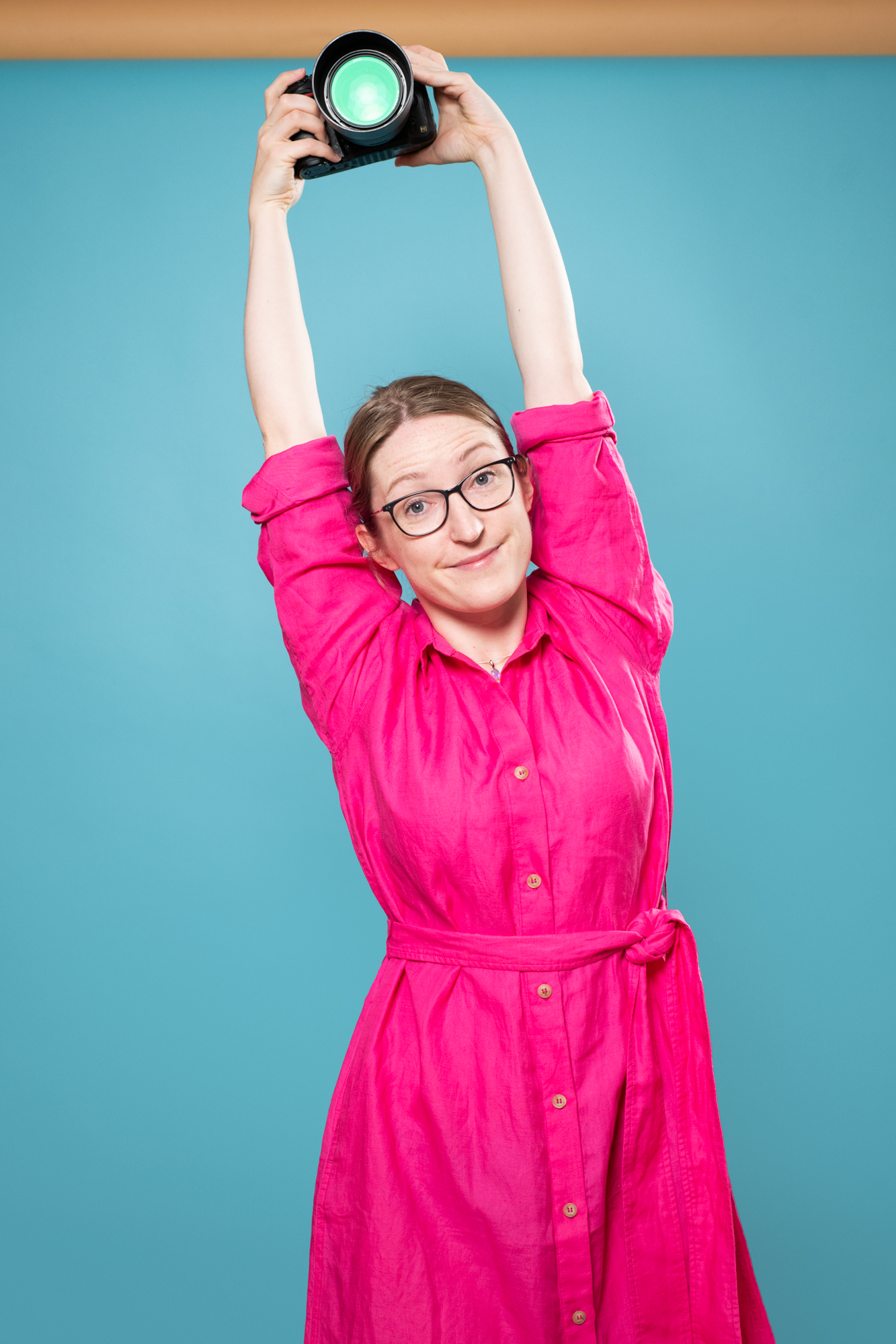 Portrait of Julia Nance in bright pink dress on a blue background holding a camera above her head, with a quirky smile.