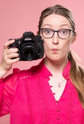 Portrait of Julia Nance on a pink background, wearing a pink shirt and holding a camera with a quirky expression (fish face).
