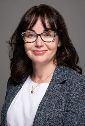 Melbourne woman smiles confidently to camera, wearing a grey blazer and patterned glasses for a relaxed but professional linkedin headshot.