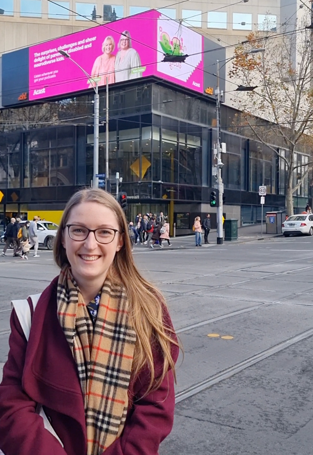 Julia stands on Bourke street smiling to camera with billboard in the background