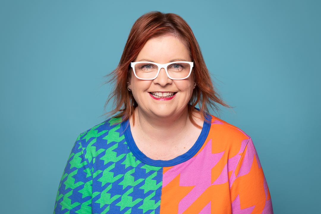 A colourful and joyful headshot of melbourne podcaster Kate, who wears a vibrant patterned jumper and white framed glasses.