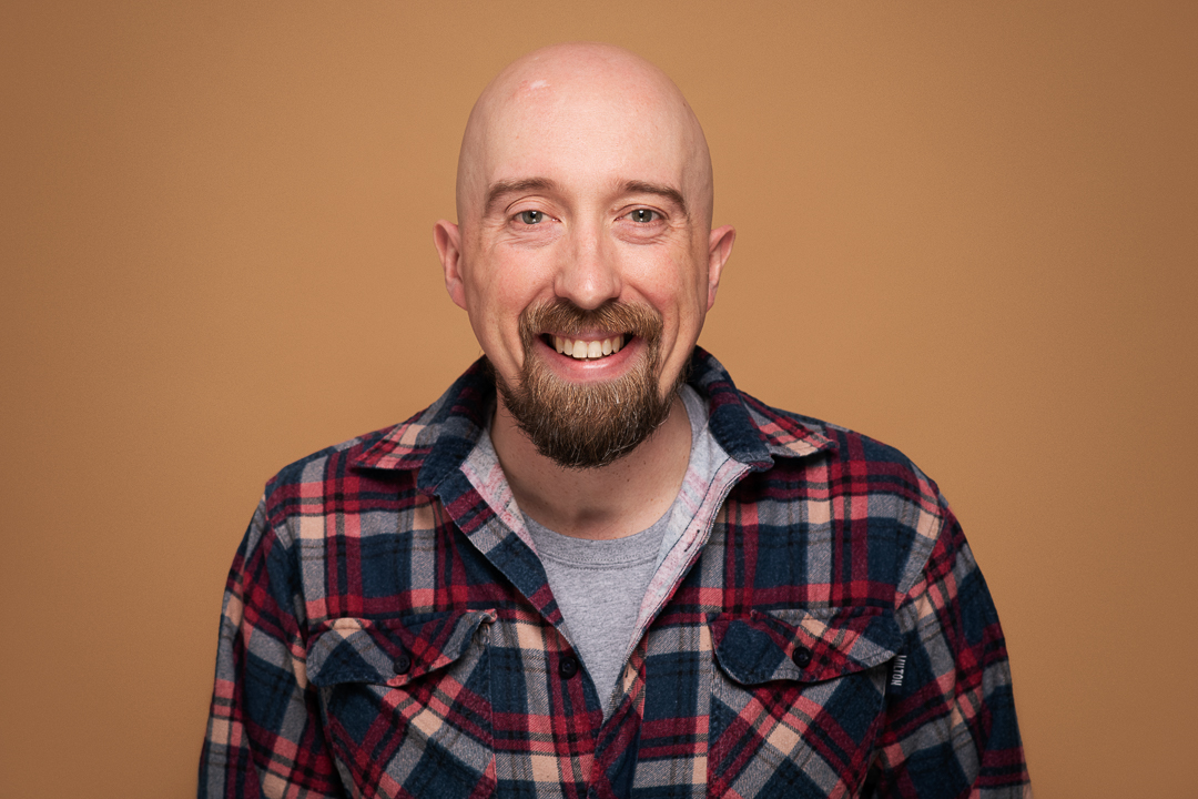 Headshot of melbourne actor Lindsay who wears a checked flannel shirt and smiles to camera on an orange background.