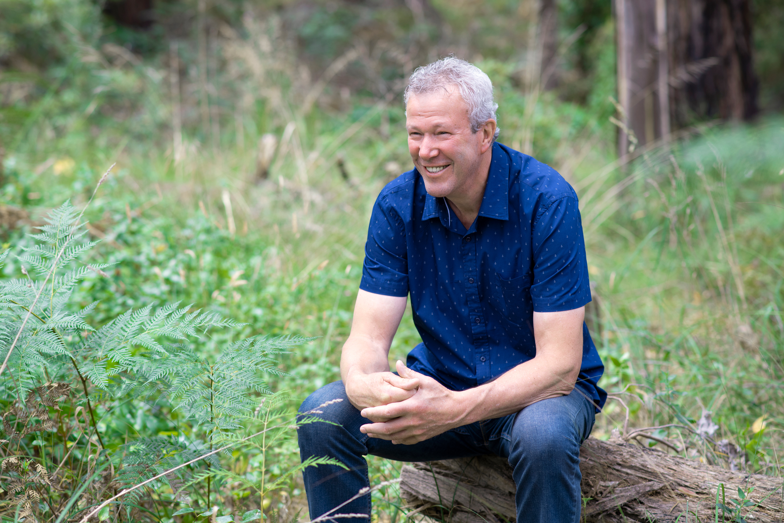 A man sits on a log for a relaxed and natural headshot for a personal branding session in Melbourne. He wears a blue shirt and jeans, and is looking away from the camera with a smile.