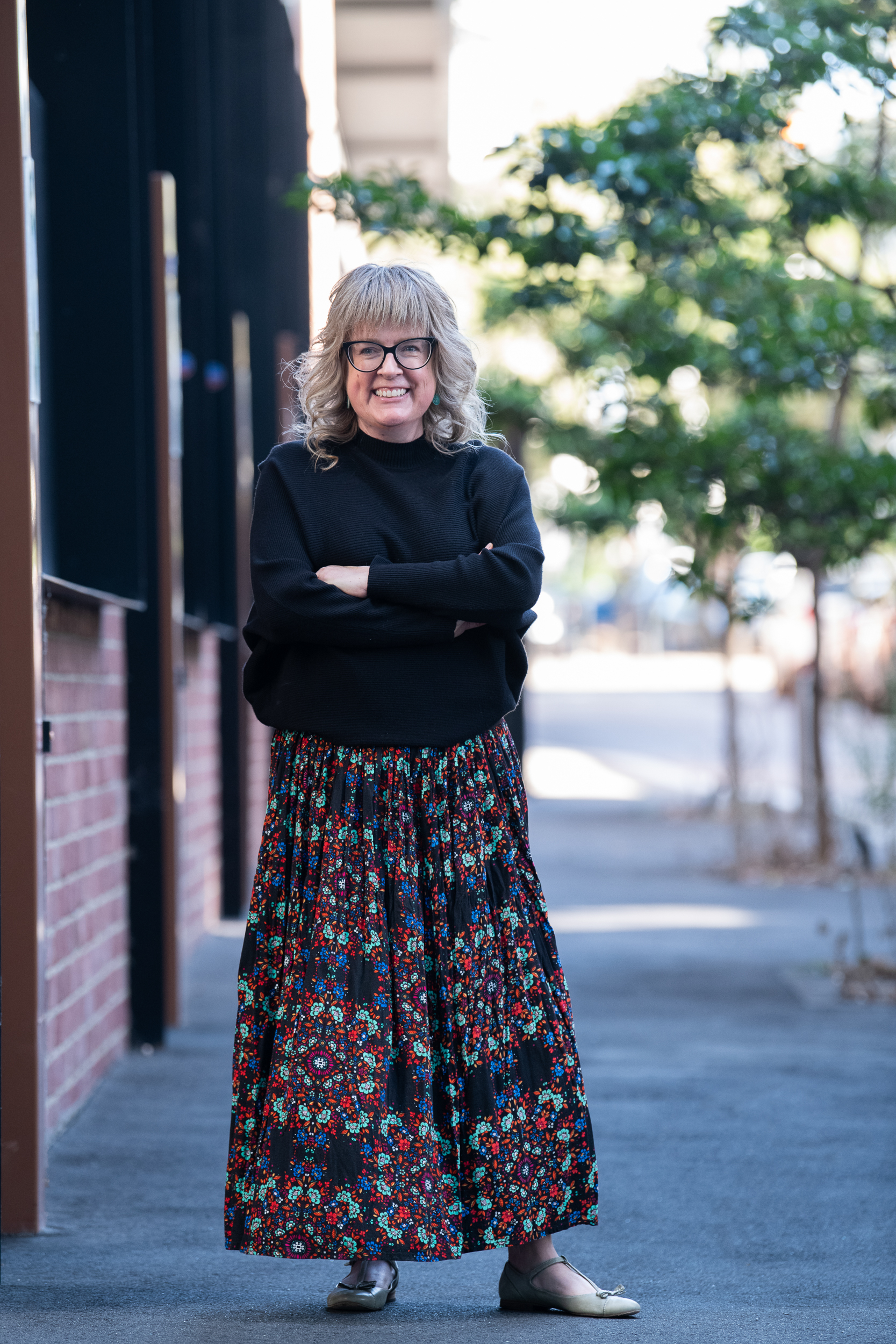 Woman stands on Melbourne street wearing black long sleeved top and patterned skirt. She has a relaxed pose and is smiling, while looking away from camera.