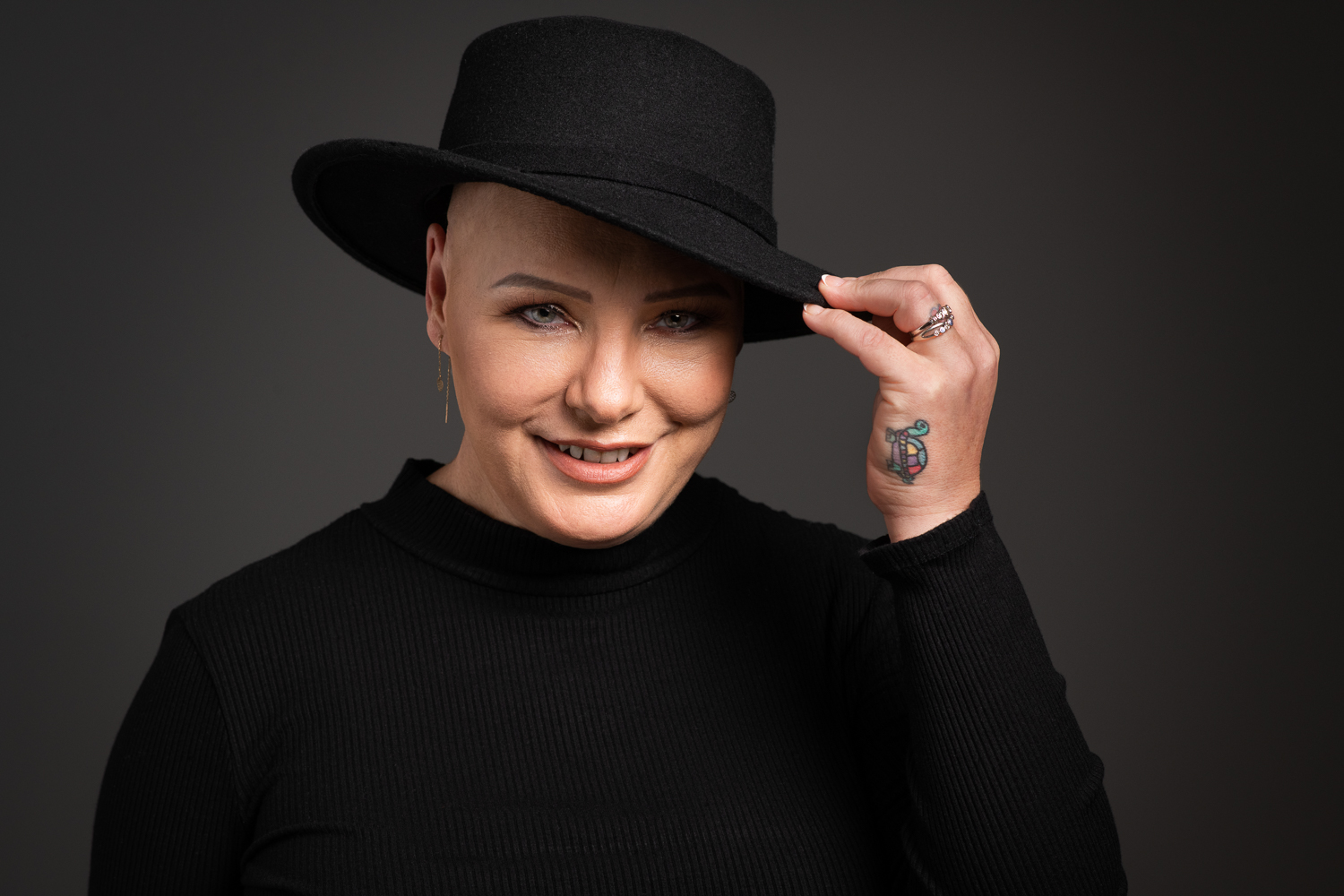 Professional portrait of woman wearing all black, smiling to camera. She has a black hat and is holding it with one hand. On her hand you can see a colourful tattoo or a turtle.